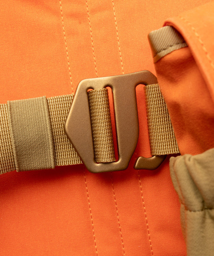 Close-up of a metal buckle securing a tan strap on orange fabric, part of the Duck Camp Canvas Upland Strap Vest.