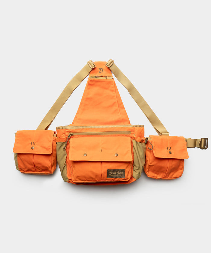 The Duck Camp Canvas Upland Strap Vest is a tan-and-orange utility chest rig with three front pouches, adjustable straps, and a central zippered compartment—ideal for hunting—shown on a plain white background.
