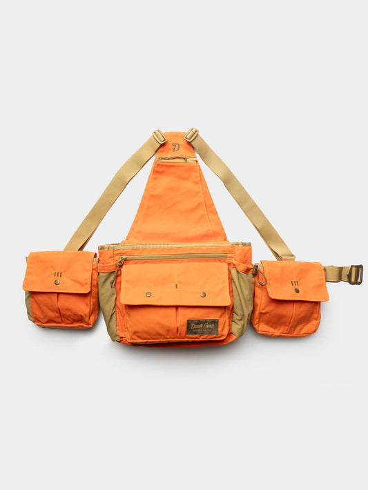 The Duck Camp Canvas Upland Strap Vest is a tan-and-orange utility chest rig with three front pouches, adjustable straps, and a central zippered compartment—ideal for hunting—shown on a plain white background.