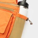 The Duck Camp Canvas Upland Strap Vest in orange and tan features a zippered pocket, field vest-inspired design, and side pocket with a white reusable water bottle with black lid.