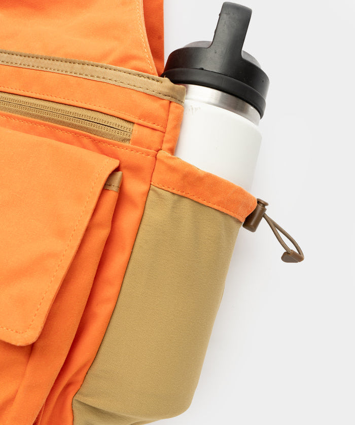Close-up of an orange and tan backpack with a white water bottle in the side pocket, shown alongside the Duck Camp Canvas Upland Strap Vest, perfect for hunting gear, against a plain white background.