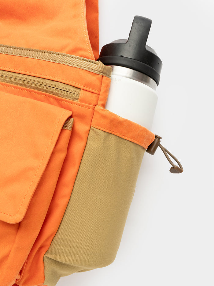 The Duck Camp Canvas Upland Strap Vest in orange and tan features a zippered pocket, field vest-inspired design, and side pocket with a white reusable water bottle with black lid.