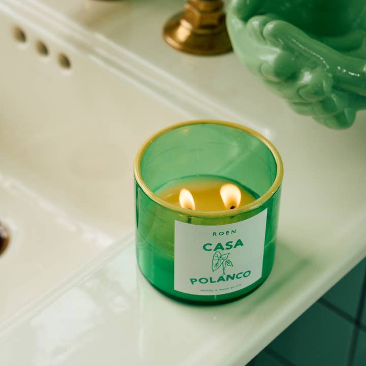 A Roen Casa Polanco | Roen double-wick hand-poured soy candle rests on a white bathroom sink next to a brass fixture and green decor.