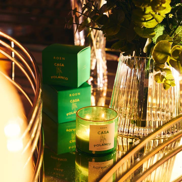 A lit green Casa Polanco | Roen candle by Roen, crafted from hand-poured soy wax, sits on a reflective surface beside its matching box and a glass vase filled with green foliage.