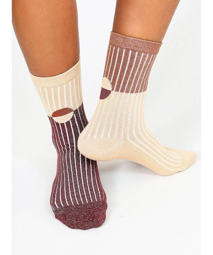 Someone wears Casey Shimmer Ankle Socks by Tailored Union: one sock has maroon and beige vertical stripes, the other beige and brown, both featuring circular accents and ribbed ankles.