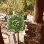 A person outdoors by a stone wall holds up the Mollyjogger CCC Legacy Bandana—100% cotton and adorned with a tree, the word "conservation," and wildlife illustrations inspired by the Civilian Conservation Corps era.