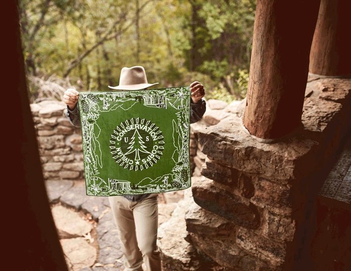 A person in a hat stands by a stone structure, holding the Mollyjogger CCC Legacy Bandana—a green 100% cotton bandana with a tree graphic and conservation message—partially concealing their face.