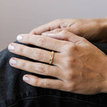A JaxKelly Gold Rings | Inset Baguette ring rests on the ring finger of a person’s left hand, which is placed on their knee, highlighting neatly manicured nails and denim jeans.