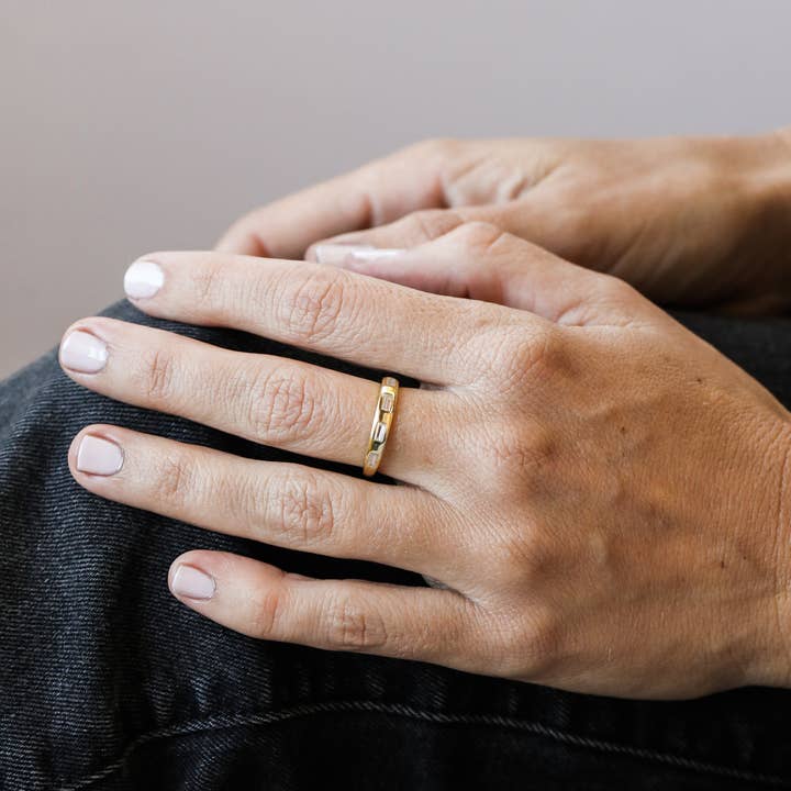 A JaxKelly Gold Rings | Inset Baguette ring rests on the ring finger of a person’s left hand, which is placed on their knee, highlighting neatly manicured nails and denim jeans.