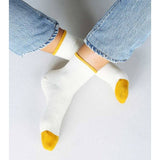 Person wears Tailored Union's Centre Ankle Sock in Off White, featuring yellow toes and heels, paired with light blue jeans. They sit cross-legged on a light background that highlights the socks’ center ankle and contrasting color details.