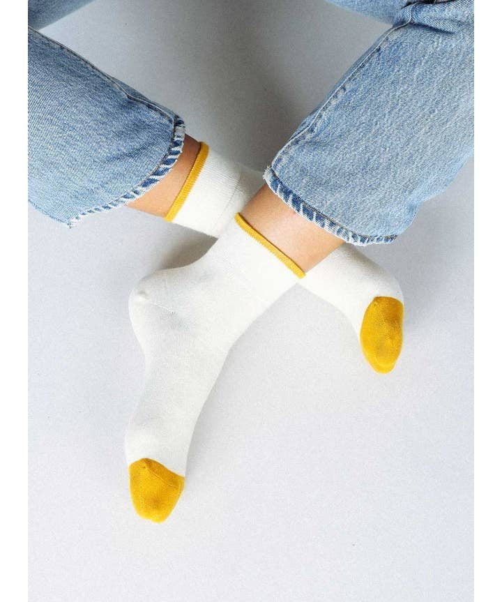 Person wears Tailored Union's Centre Ankle Sock in Off White, featuring yellow toes and heels, paired with light blue jeans. They sit cross-legged on a light background that highlights the socks’ center ankle and contrasting color details.