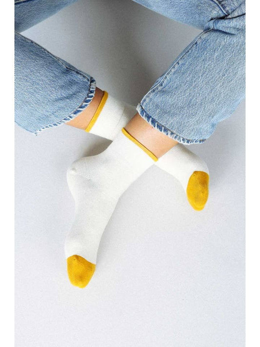 A person sits cross-legged on a light background, wearing light blue jeans and Tailored Union Centre Ankle Sock–Off White with yellow toes and cuffs, exposing the socks at the ankles.