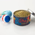 An open tin of Beautiful Briny Sea's Cha Cha Delish Seasoning, a citrus spice blend, rests next to two colorful maracas on a white surface.