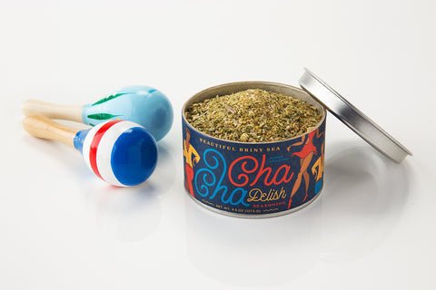 An open tin of Beautiful Briny Sea's Cha Cha Delish Seasoning sits beside two blue, white, and red maracas on a white surface.