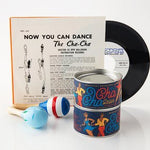 A cha-cha dance record, its cover with dance steps and maracas, sits beside a decorative tin of Cha Cha Delish Seasoning by Beautiful Briny Sea—each artfully arranged on a white surface.