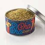 An open tin of Cha Cha Delish Seasoning by Beautiful Briny Sea sits on a white surface, its lid placed beside it.