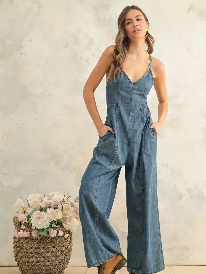 A woman models the Chambray Fabric Jumpsuit by Miou Muse, standing beside a woven basket filled with light-colored flowers against a neutral textured wall.