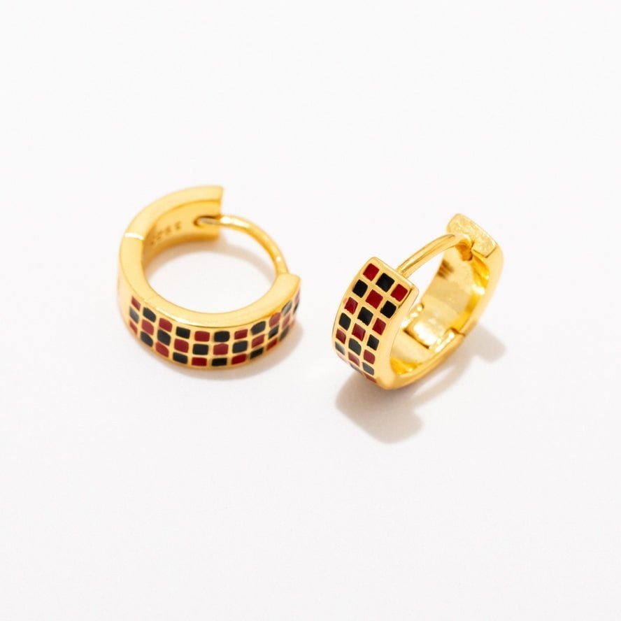 The Checker Huggie Hoops by Larissa Loden are 18k gold plated, hypoallergenic earrings featuring a pattern of small red and black squares on a white background—perfect for comfortable everyday wear.