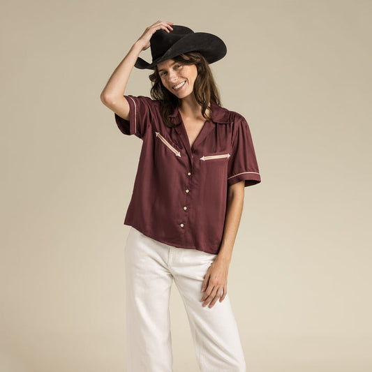 A woman wearing a maroon Cheyenne Western Shirt by Sendero Provisions Co., black cowboy hat, and white pants smiles against a plain beige background.