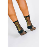 A person models Tailored Union’s Chic Sheer Ankle Sock—black, sheer, cotton-nylon blend with a vertical yellow and black checkered stripe at the back—while standing against a white background.