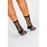 A person wearing Tailored Union’s Chic Sheer Ankle Sock—black sheer cotton nylons with a vertical yellow and black checkered stripe on the back—stands on a white background.