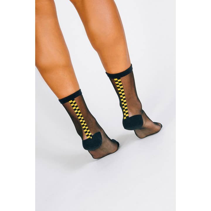 A person wearing Tailored Union’s Chic Sheer Ankle Sock—black sheer cotton nylons with a vertical yellow and black checkered stripe on the back—stands on a white background.