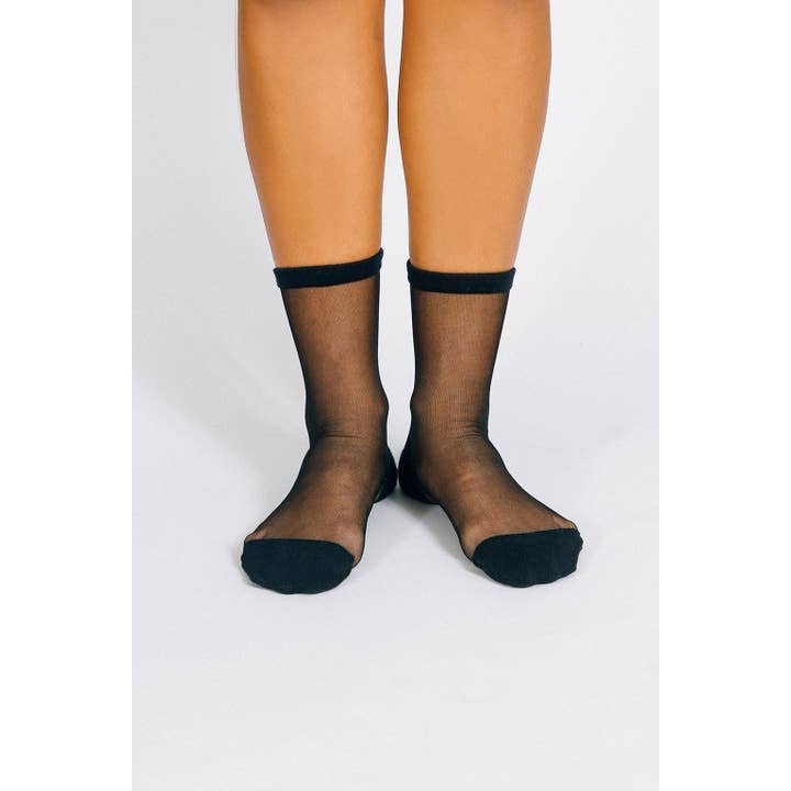 A person wearing Tailored Union’s Chic Sheer Ankle Sock stands on a white surface, highlighting the stylish contrast of its sheer black design with solid black toes, heels, and cuffs.