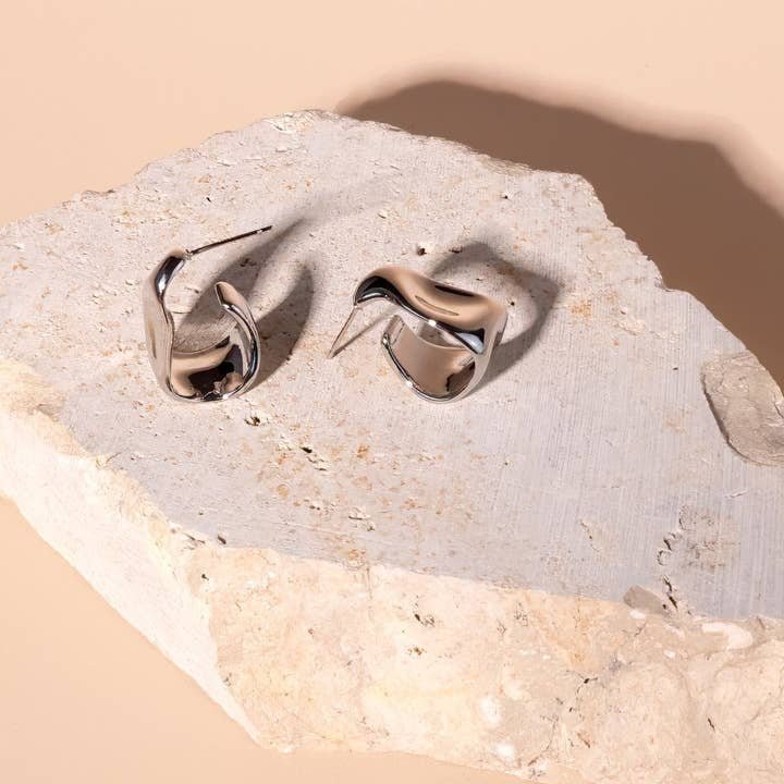 The Chloe Hoops by Peter and June—modern, abstract silver rhodium plated earrings—are displayed on a rough light stone with a beige background.