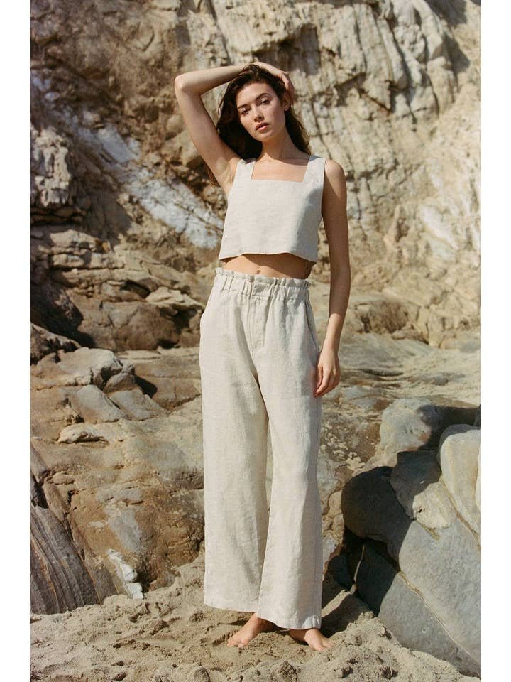 A woman stands barefoot on rocky terrain, wearing a sleeveless beige crop top and the Christal Pants by A. Ren—a pair of high-waisted linen pants featuring a ruffled waist.
