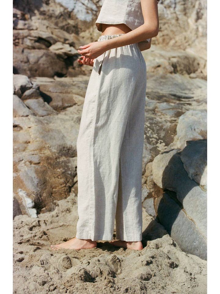 A person stands barefoot on sandy rocks at the beach wearing the Christal Pants by A. Ren—loose linen pants with a ruffled waist—paired with a sleeveless crop top.