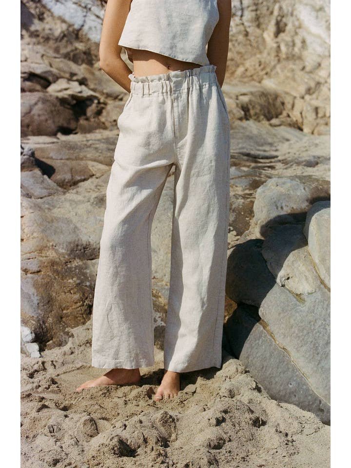 A person stands barefoot on sand and rocks, wearing the Christal Pants by A. Ren—loose, light-colored linen pants with a ruffled waist—paired with a matching sleeveless top. Their head and upper torso are not visible.