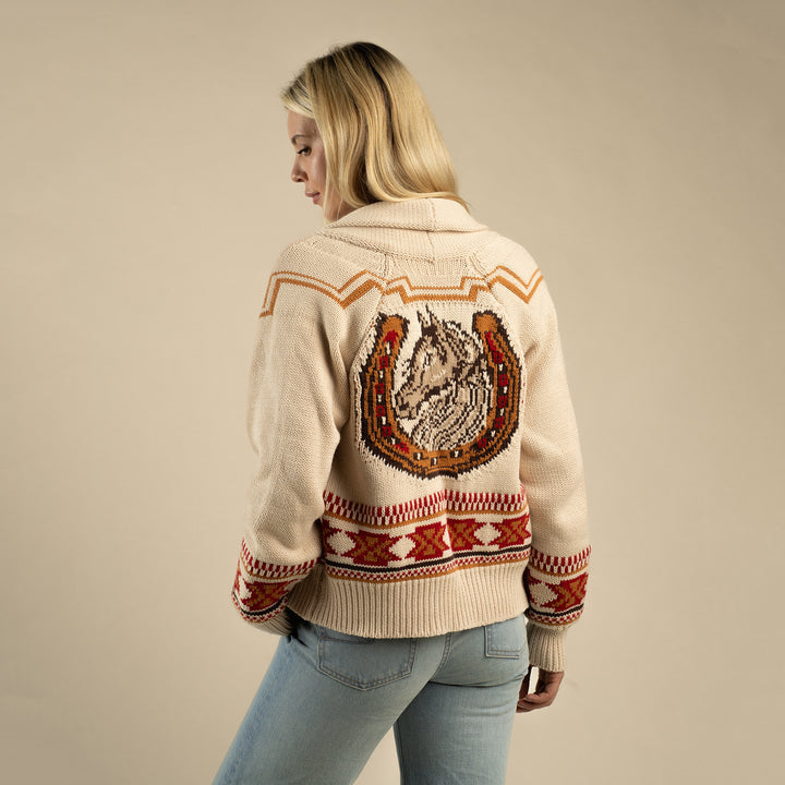 A woman faces away in the Sendero Provisions Co. Cimarron Cardigan—a beige knit with a large horseshoe and horse design on the back, Western details, and bold red geometric patterns on the hem and sleeves.