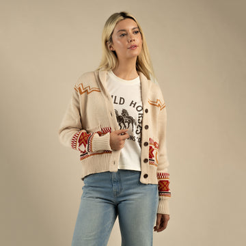 A woman stands against a plain background wearing the Cimarron Cardigan by Sendero Provisions Co. over a white graphic tee and blue jeans, effortlessly channeling Southwest style.