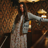 A woman in a floral dress and the Cimarron Cardigan by Sendero Provisions Co. stands indoors by a wooden railing, with ornate wallpaper, Western accents, and a mirror in the background.
