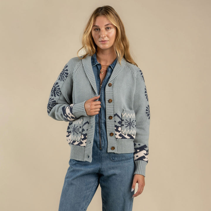 A woman stands against a beige background wearing the blue patterned Cimarron Cardigan by Sendero Provisions Co. over a denim shirt and jeans.