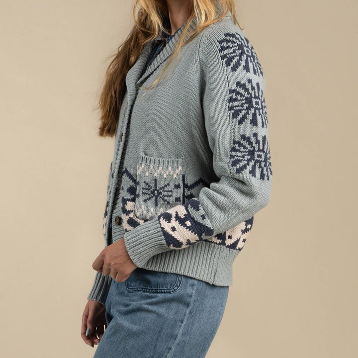 A person wears the Cimarron Cardigan by Sendero Provisions Co.—a light blue knit with dark blue and white snowflake patterns, subtle Western details, and pairing it with blue jeans against a beige background for a cozy, casual style.