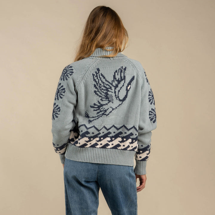 A person faces away, wearing Sendero Provisions Co.'s Cimarron Cardigan in light blue with a large bird design and Western details on the sleeves and waistband, paired with blue pants.