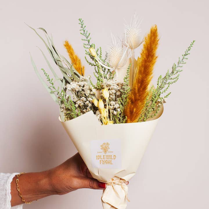 A hand holds the Citrine Dried Flower Bouquet by Idlewild Floral Co., featuring assorted dried grasses and blooms wrapped in cream-colored paper with the Idlewild Floral Co. label.
