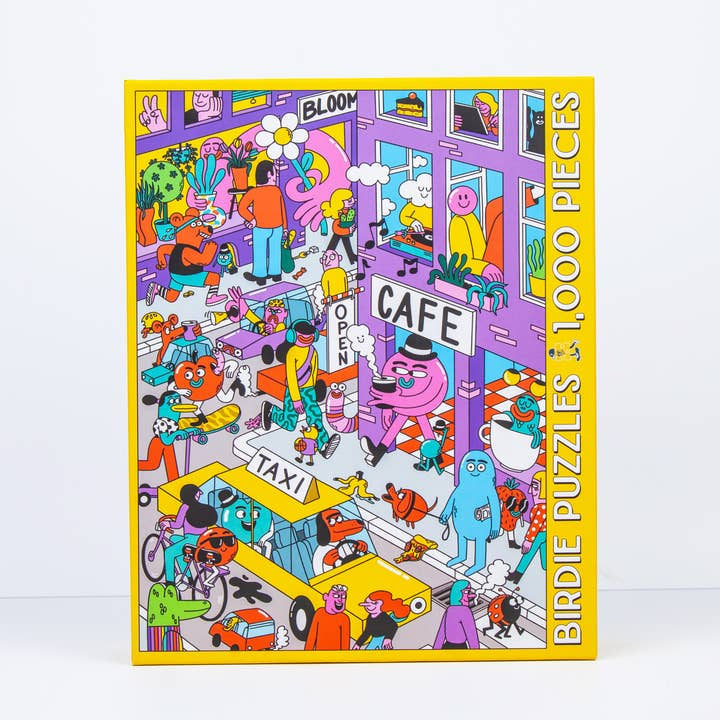 The City Vibes puzzle by Birdie Puzzles features a colorful cityscape by Sarah Matuszewski with people, animals, a taxi, and café. The 1,000-piece box is made from recycled materials and displays “Birdie Puzzles” along the side.