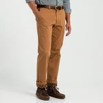 Man wearing tan Classic Fit Brushed Back Chino by Duck Head, brown leather belt, checkered shirt, and brown boat shoes stands with hands in pockets against a plain background.