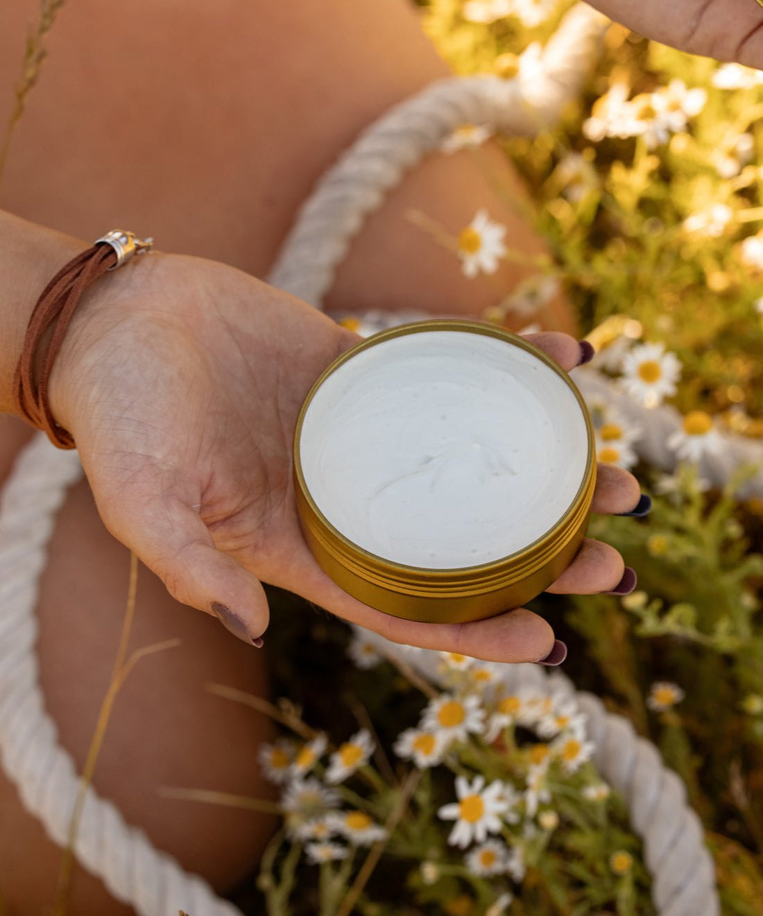 Someone holds an open tin of COASTAL COWGIRL Cocoa and Coconut Tallow Body Butter by Cowboy & Co. while relaxing among grass and small white flowers outdoors.