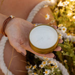 Someone holds an open tin of COASTAL COWGIRL Cocoa and Coconut Tallow Body Butter by Cowboy & Co. while relaxing among grass and small white flowers outdoors.