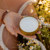 Someone holds an open tin of COASTAL COWGIRL Cocoa and Coconut Tallow Body Butter by Cowboy & Co. while relaxing among grass and small white flowers outdoors.