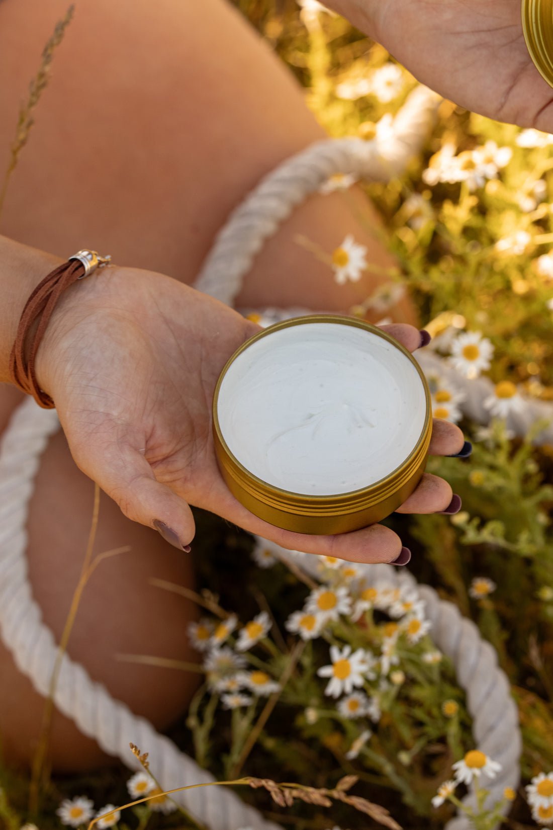 Someone holds an open tin of COASTAL COWGIRL Cocoa and Coconut Tallow Body Butter by Cowboy & Co. while relaxing among grass and small white flowers outdoors.