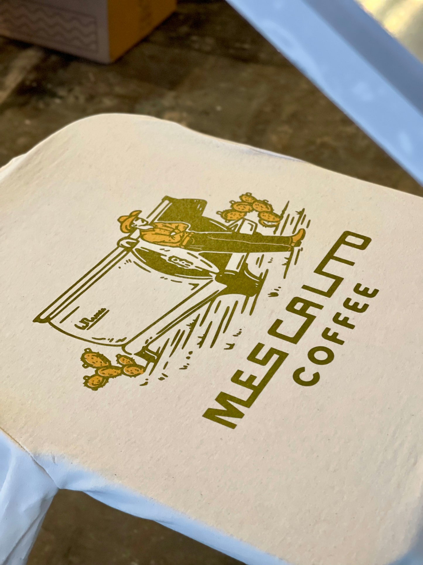 A printed fabric featuring a graphic of a cowboy beside a cart and the text "MES CALTO COFFEE"—celebrating western style, inspired by the Coffee Cowboy | Mescalito Coffee shirt from Mescalito Coffee.