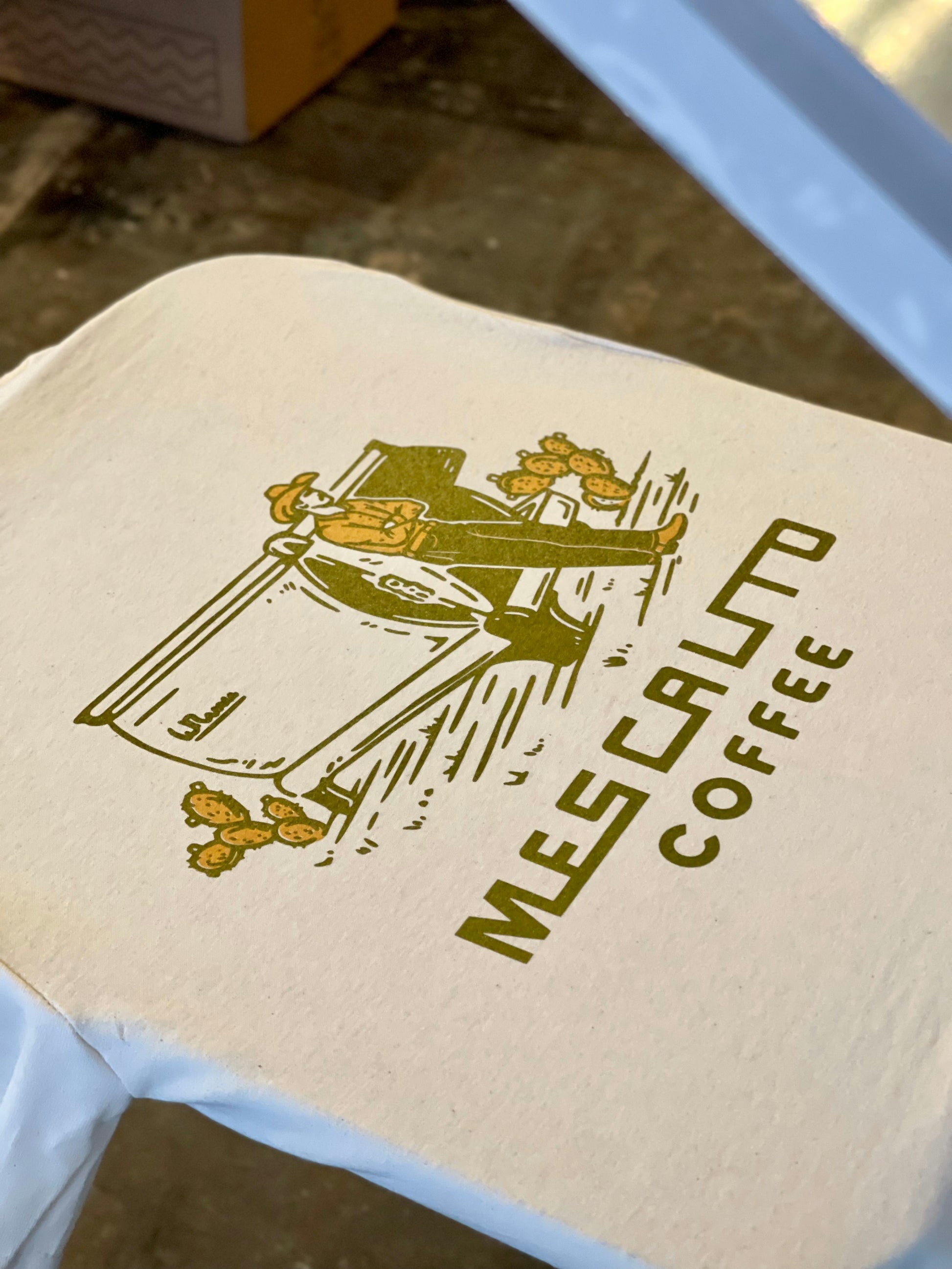 A printed fabric featuring a graphic of a cowboy beside a cart and the text "MES CALTO COFFEE"—celebrating western style, inspired by the Coffee Cowboy | Mescalito Coffee shirt from Mescalito Coffee.