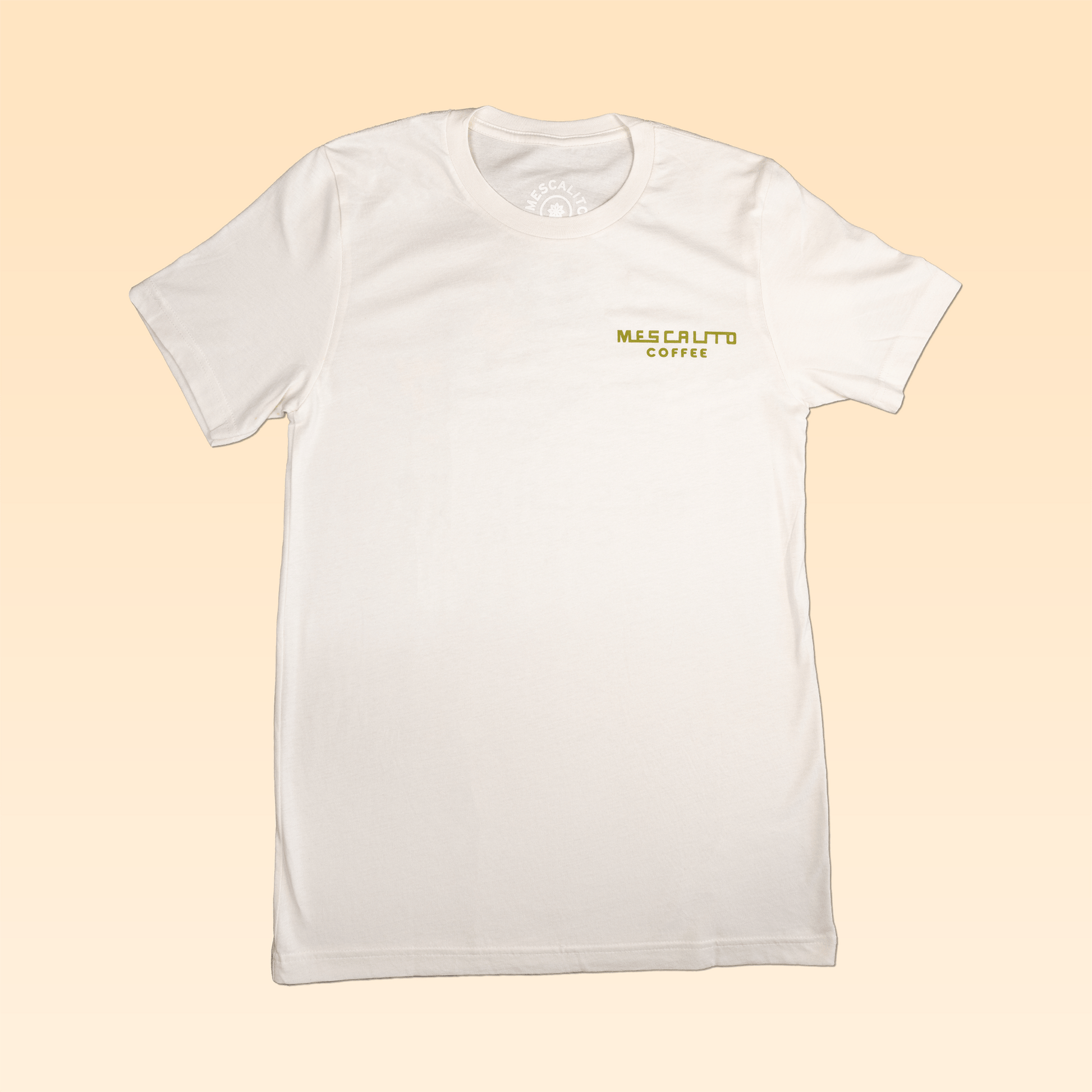 A plain white Coffee Cowboy | Mescalito Coffee shirt by Mescalito Coffee with "MEZCALITO COFFEE" in yellow and green on the upper left chest, shown against a light beige background.
