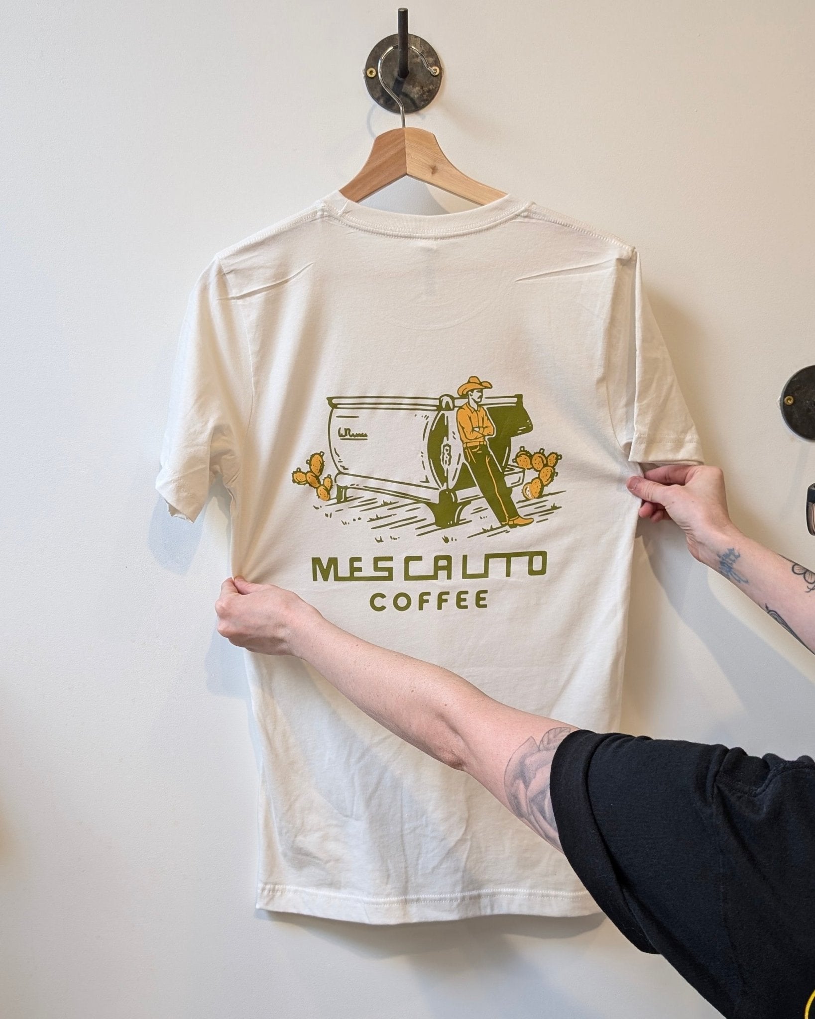 A person holds up a white Coffee Cowboy | Mescalito Coffee T-shirt on a hanger, featuring a farmer, coffee plants, and “Mesa Alto Coffee” text—a unique Mescalito Coffee shirt inspired by cowboy culture.