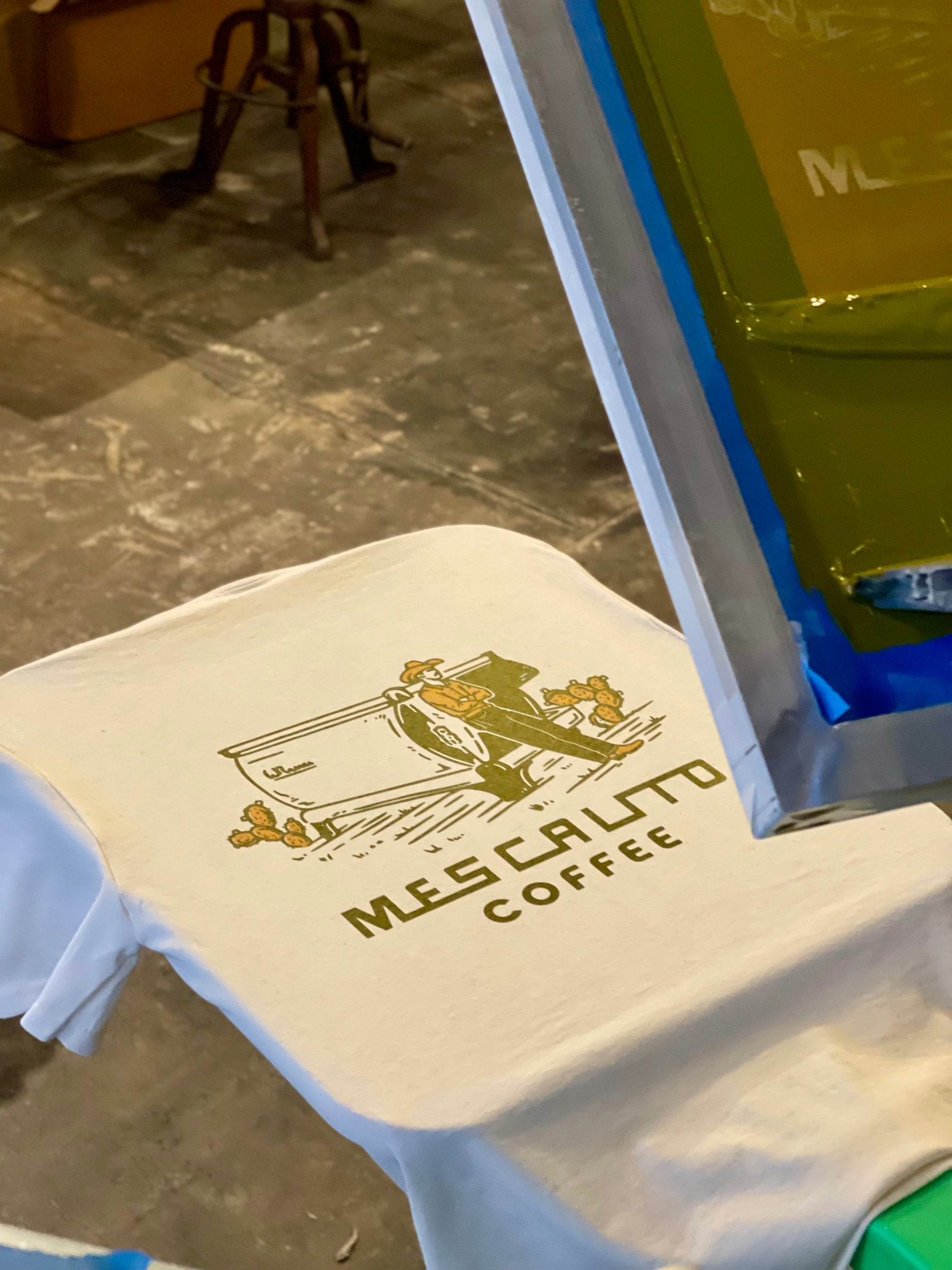 A white tee by Mescalito Coffee features the "Coffee Cowboy | Mescalito Coffee" design, capturing cowboy culture with a person and cacti in front of a camper.