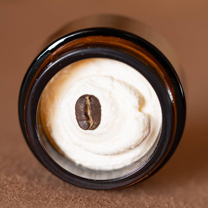 A top-down view of the Cowboy & Co. Coffee/Vanilla Infused Tallow Brightening Eye Cream shows a white cream in a jar, topped with a coffee bean.